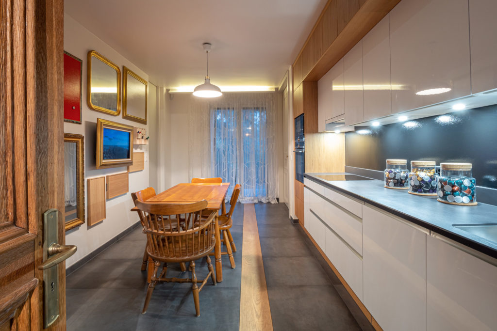 Aménagement intérieur haut de gamme à Genève : cuisine en longueur avec meubles blancs, plan de travail sombre, table en bois et chaises, cadres dorés au mur, appartement La Gradelle par le studio Camille Brunot