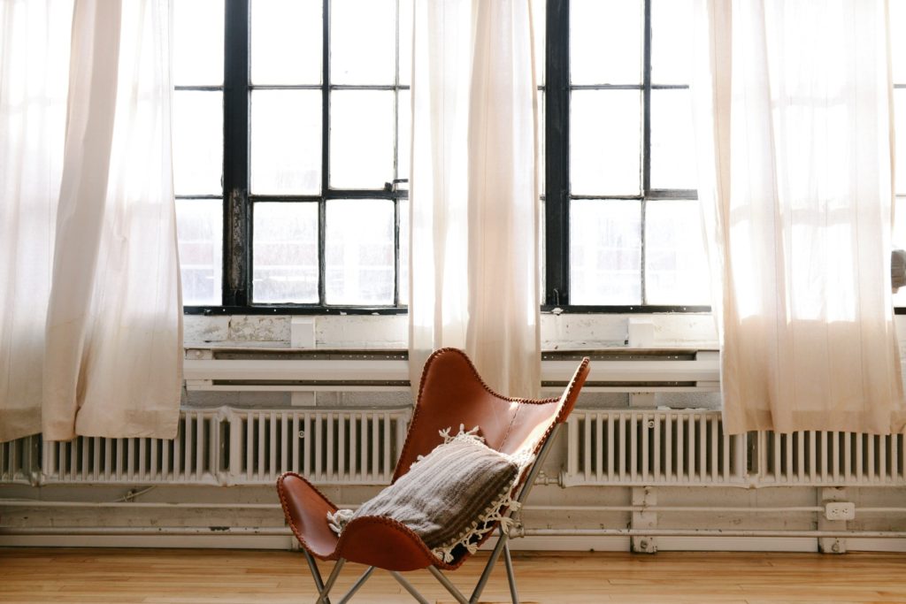 Voilages blancs légers devant de grandes fenêtres à petits carreaux, fauteuil en cuir et parquet en bois, illustration de rideaux sur mesure à Genève